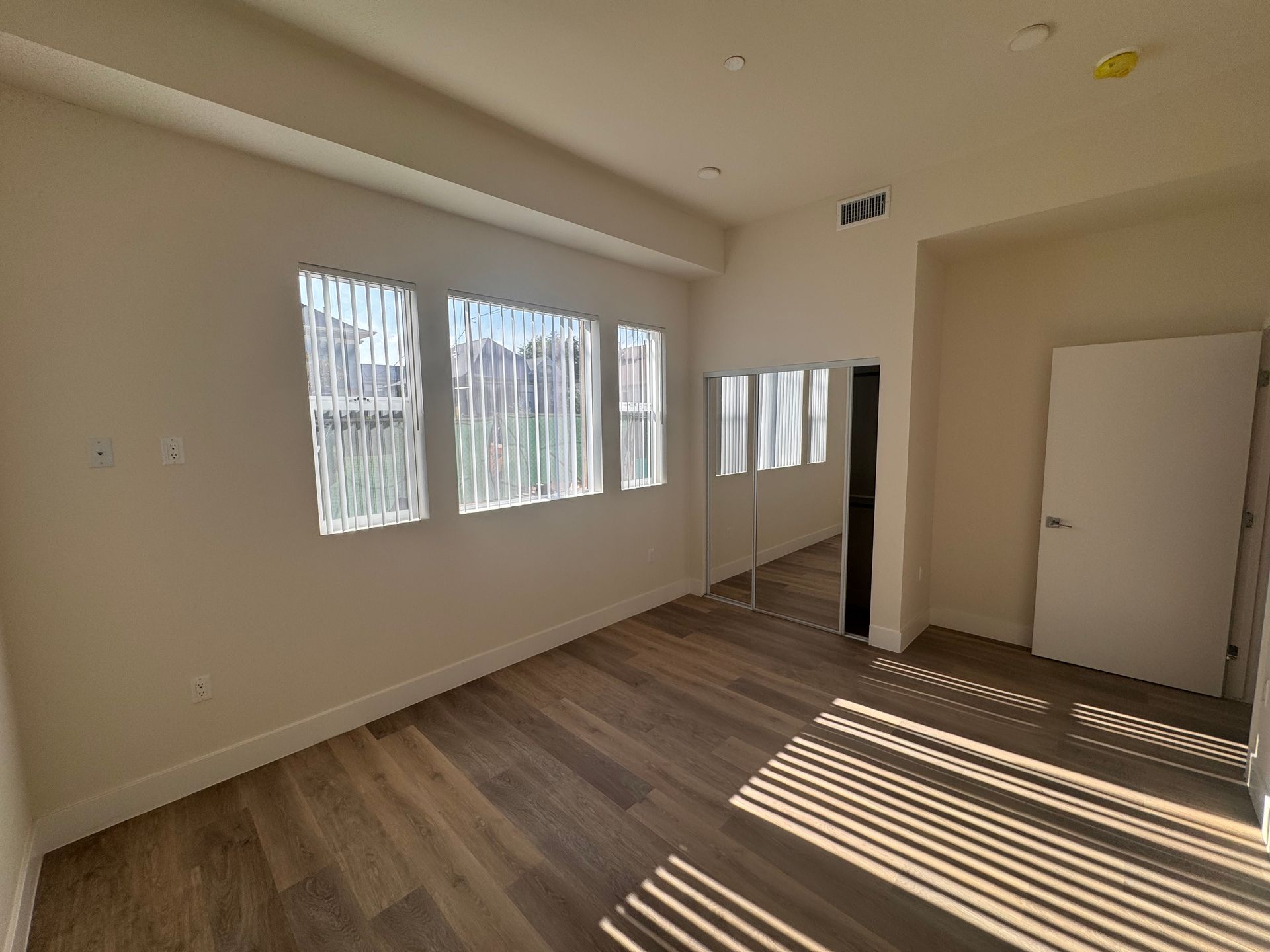 An empty room with light wood flooring, beige walls, three windows with vertical blinds, a mirrored closet, and a door.