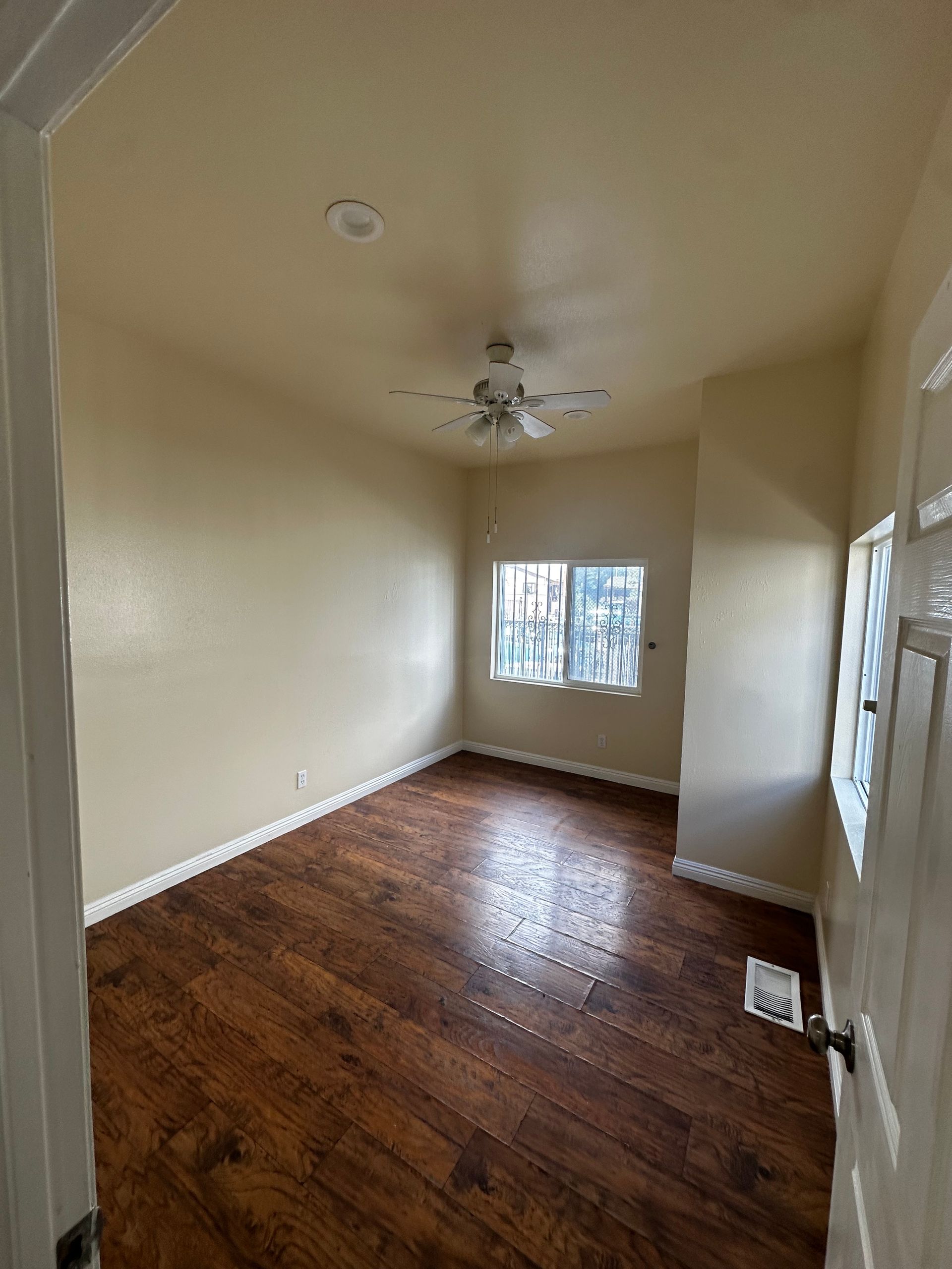 An empty room with tan walls, dark wood-look flooring, a ceiling fan, and windows on two walls.