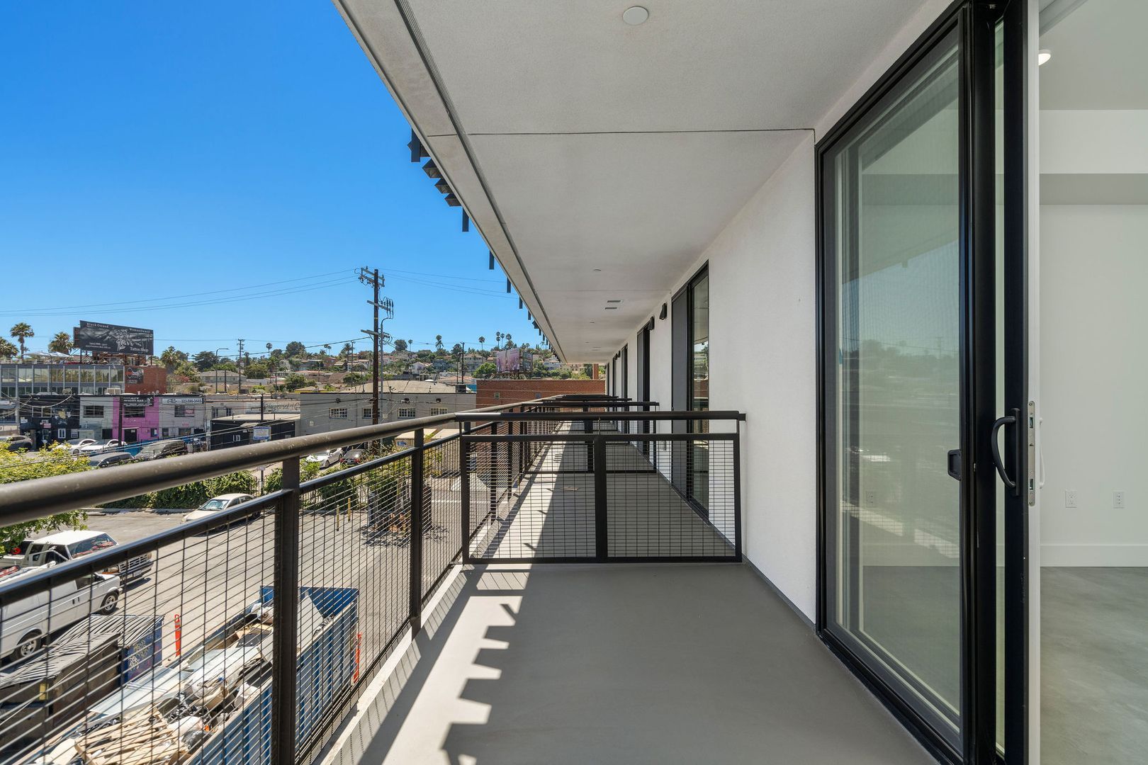 A covered balcony with a black railing, looking out over a sunlit urban landscape with distant buildings.