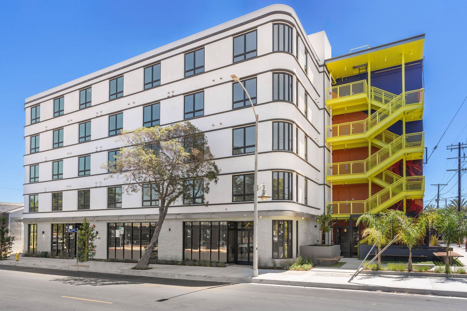 A modern, five-story white building with rounded corners, ground-floor retail, and a bright yellow outdoor stairwell.