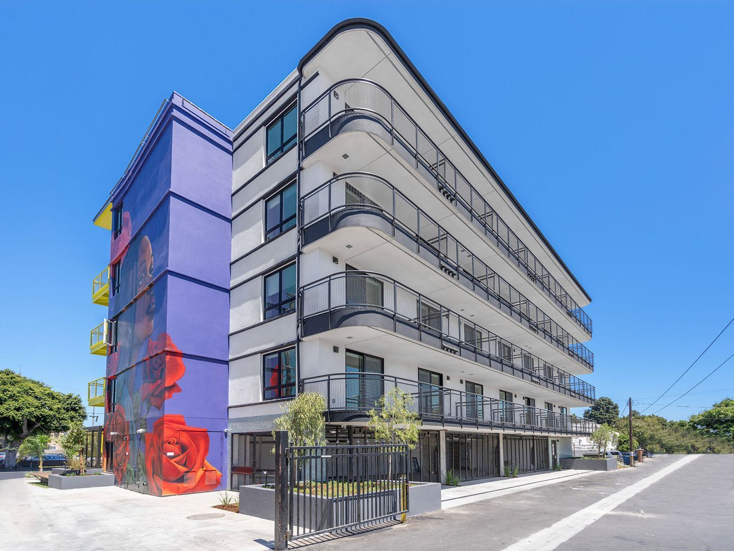 A modern five-story apartment building with a white facade, black railings, and a purple pillar painted with red roses.