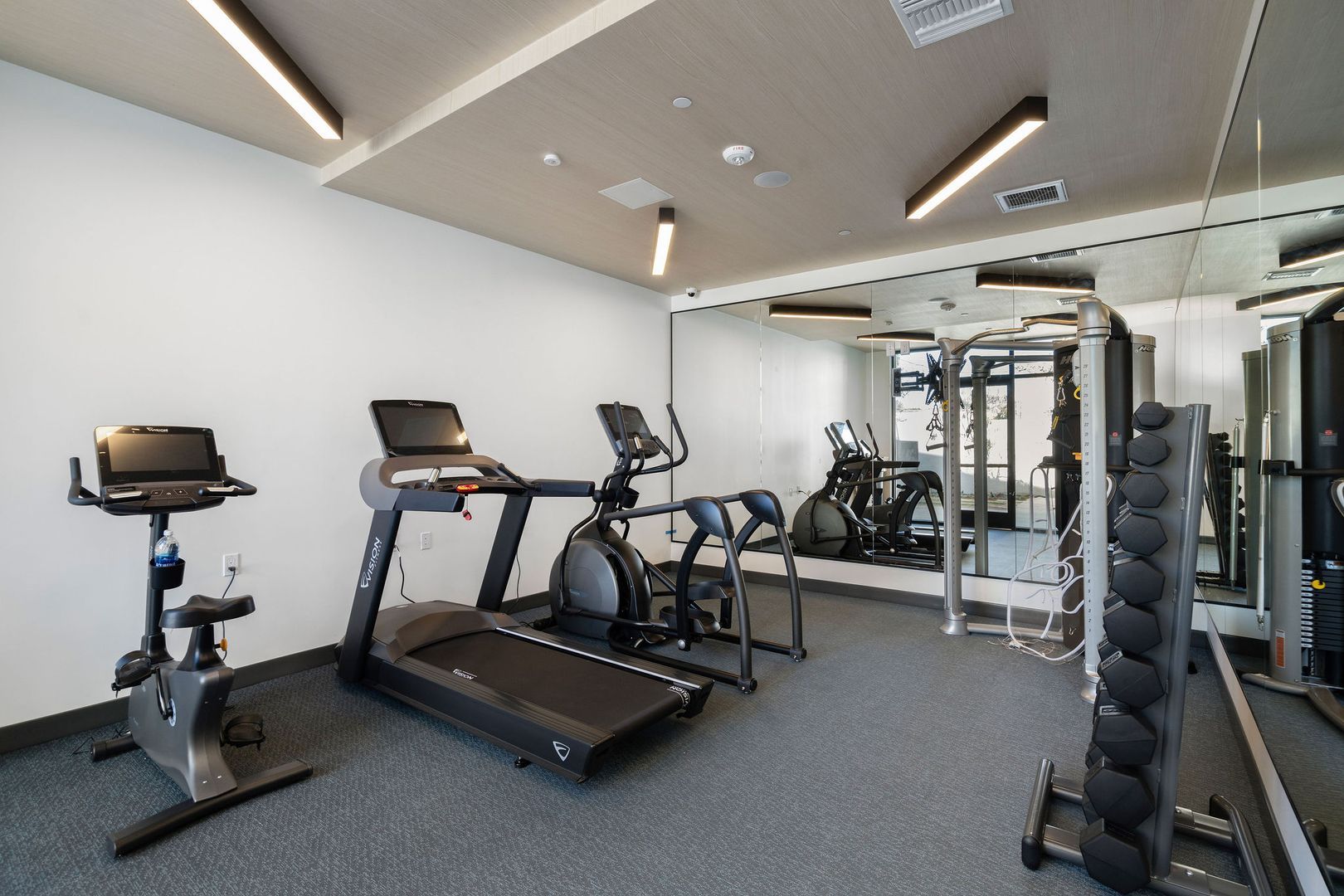 A compact gym featuring a stationary bike, treadmill, elliptical, and a rack of dumbbells against a mirrored wall.
