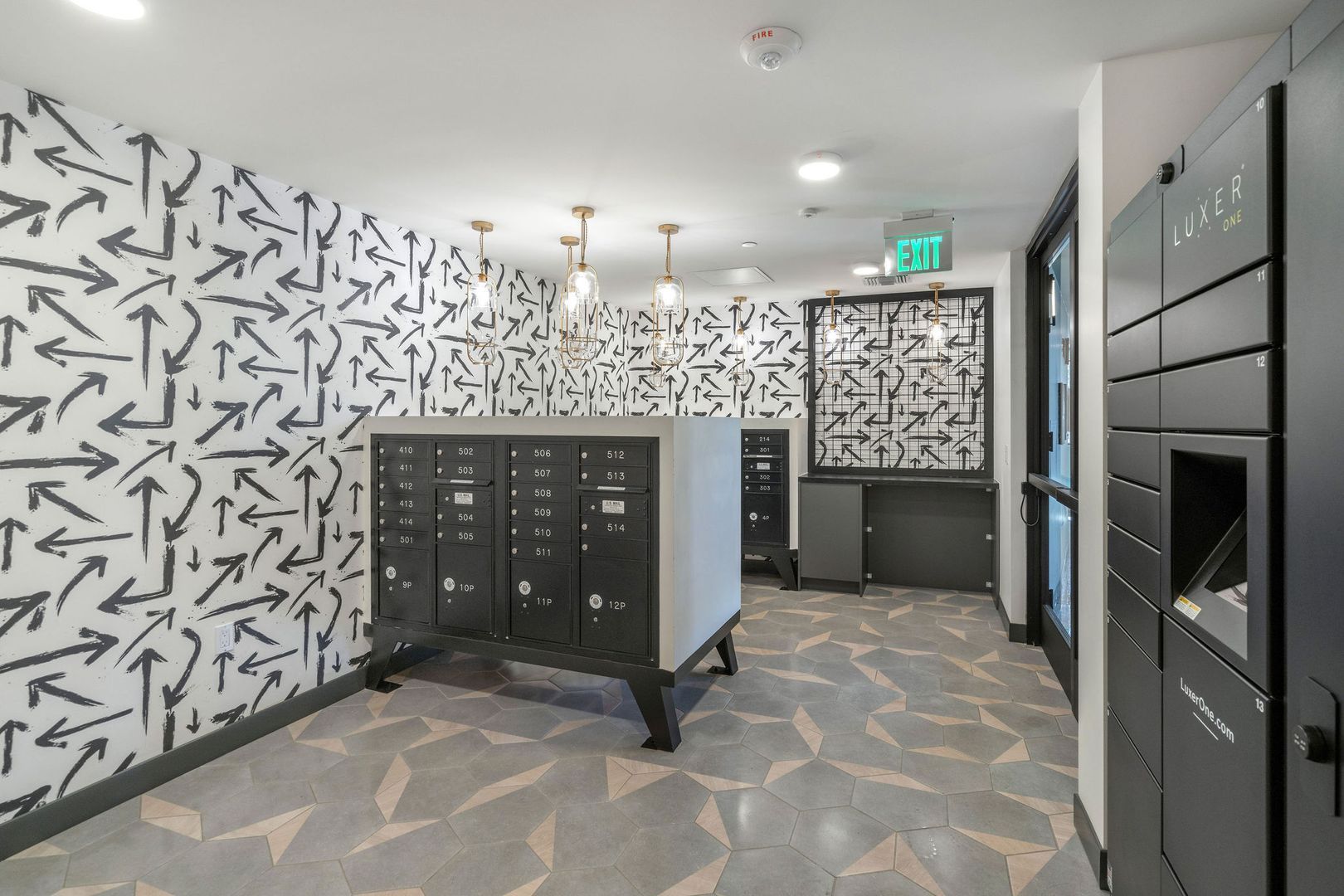 A mail room with black locker banks, patterned arrow wallpaper, and grey tiled flooring, featuring an exit sign above.