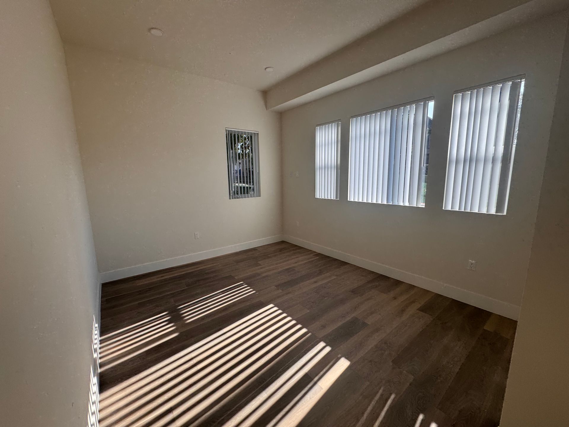 A sunlit, empty room with dark wood flooring, beige walls, and three windows covered with white vertical blinds.