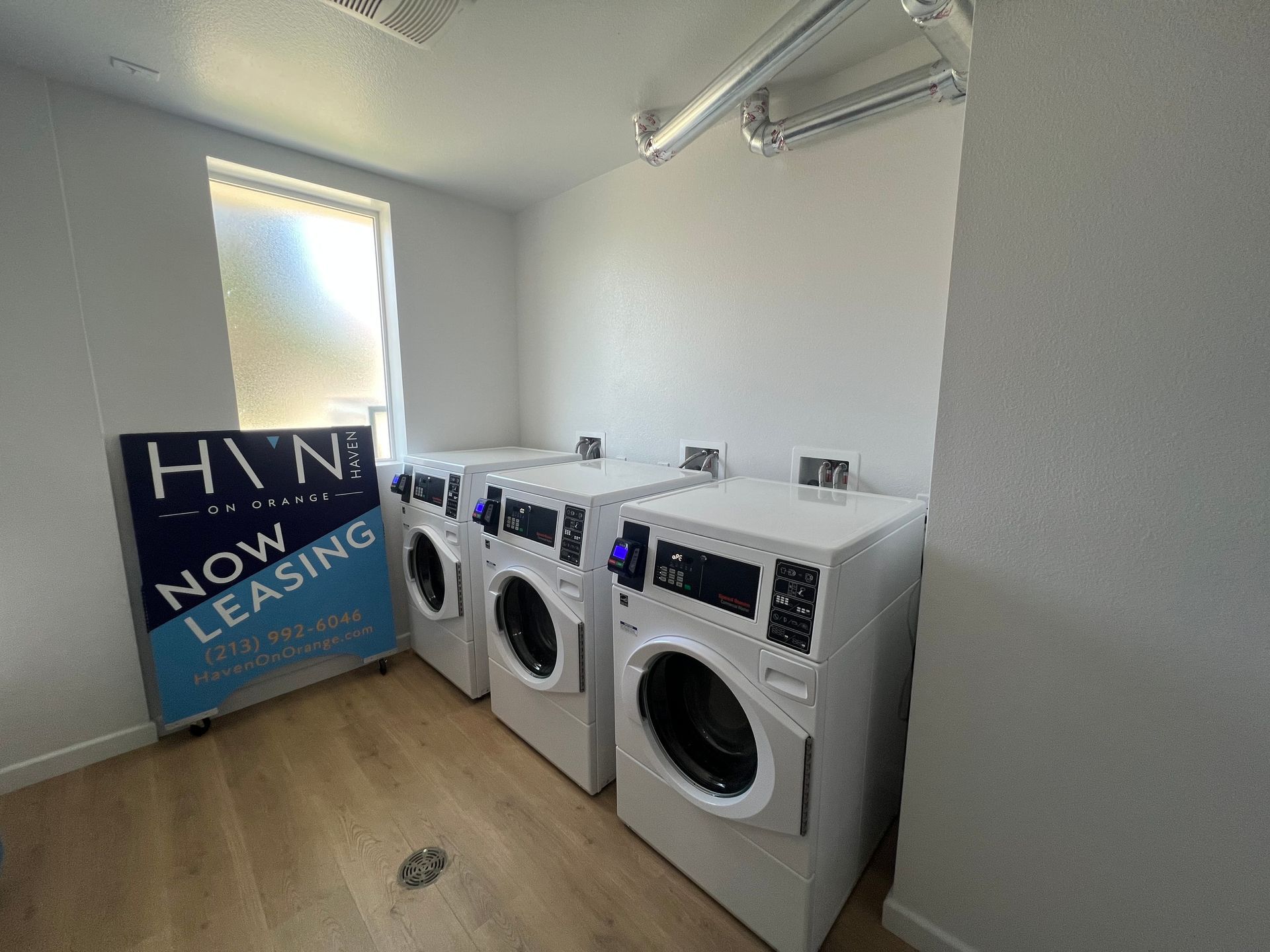 A laundry room with three white washers next to a leasing sign near a window.