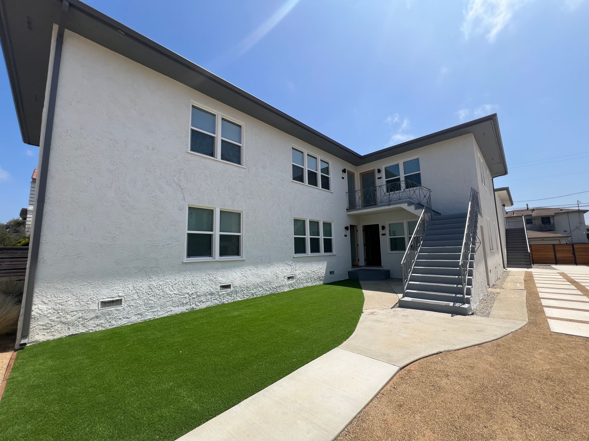 A two-story white building with gray trim, a staircase leading to a second-floor entrance, a lawn, and a stone walkway.