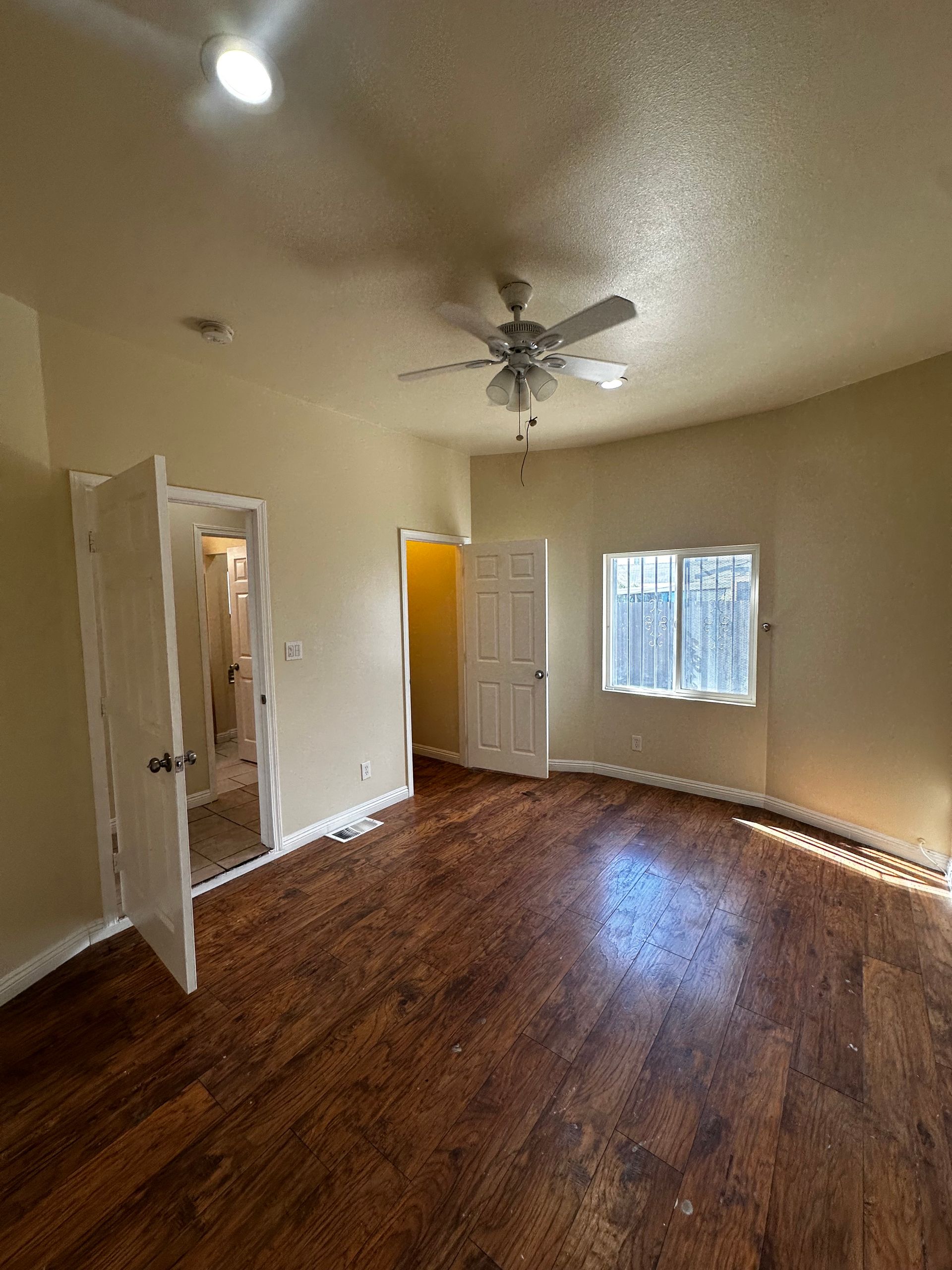 An empty bedroom with wood-look flooring, beige walls, a ceiling fan, a window, and two open white doors.