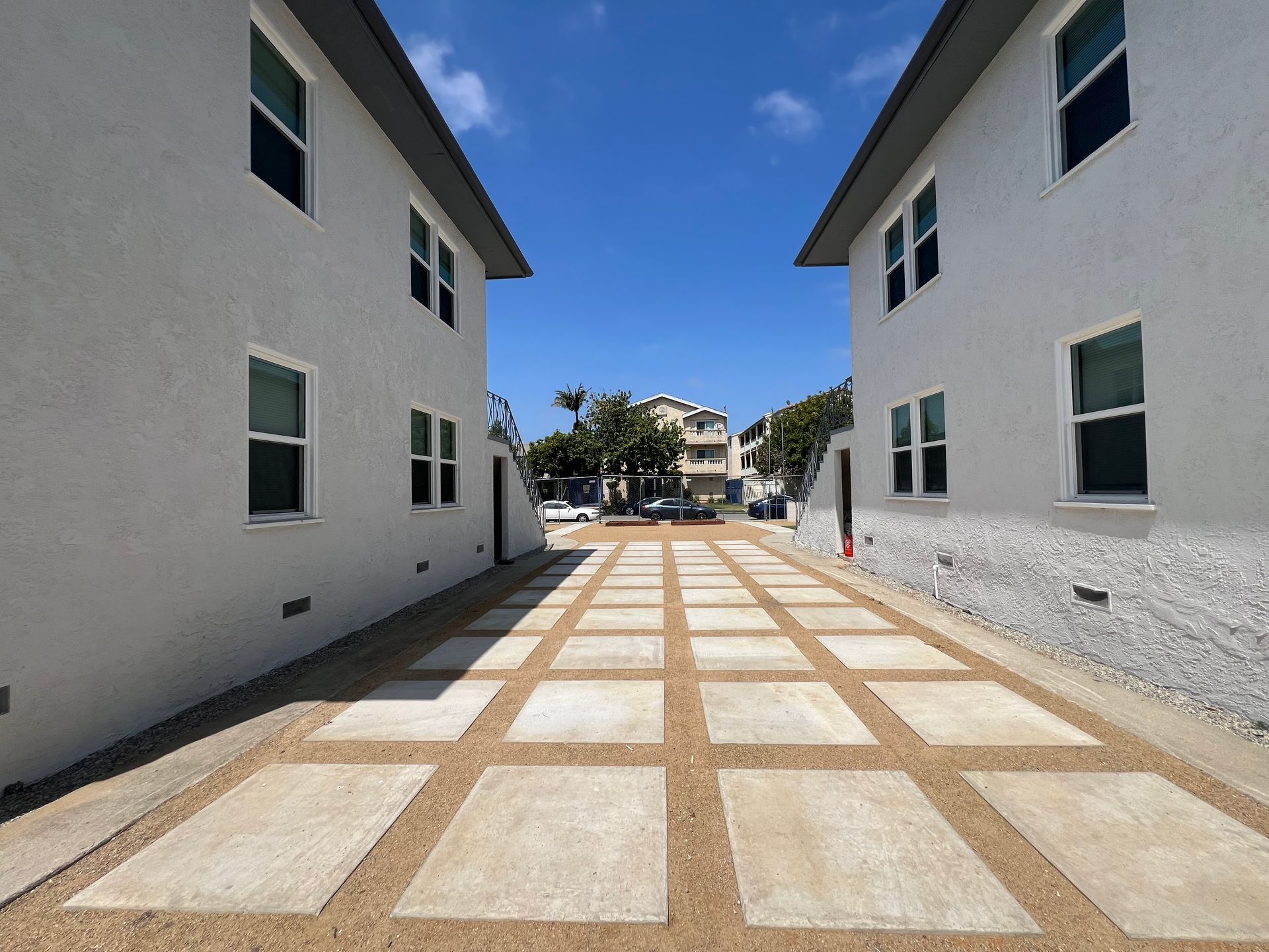 A paved outdoor walkway between two white multi-story buildings, leading to a street with parked cars and trees.