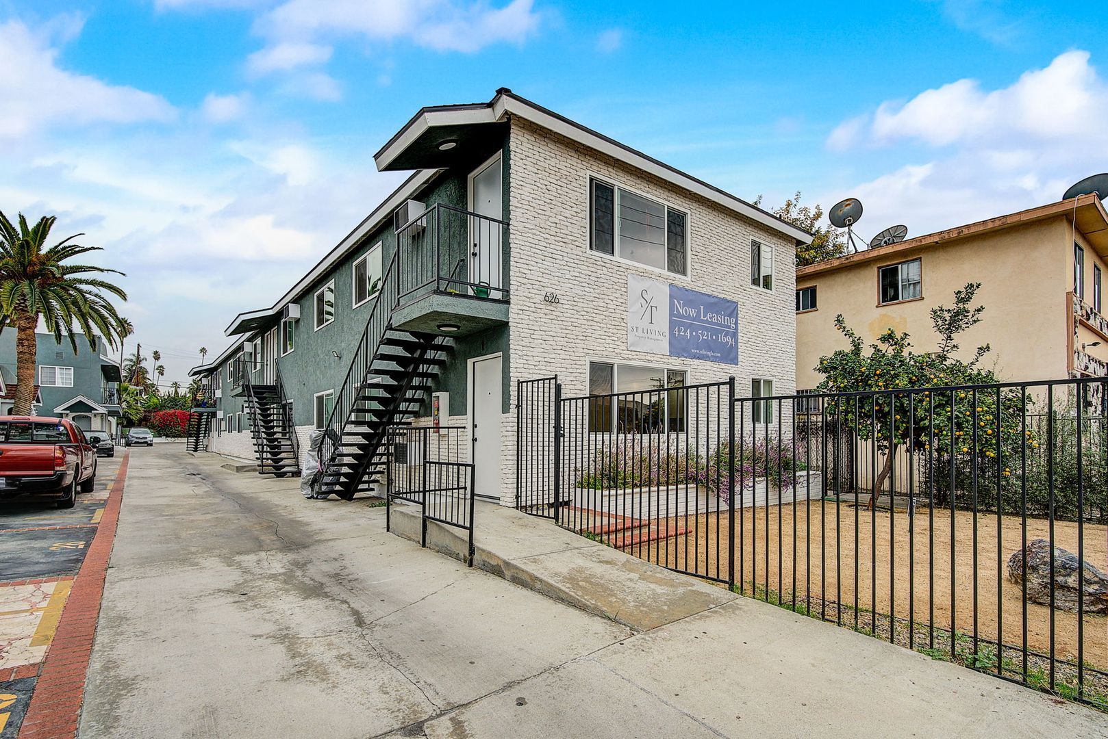 Two-story apartment building with a white brick facade and dark trim next to a paved driveway and a fenced-in yard.