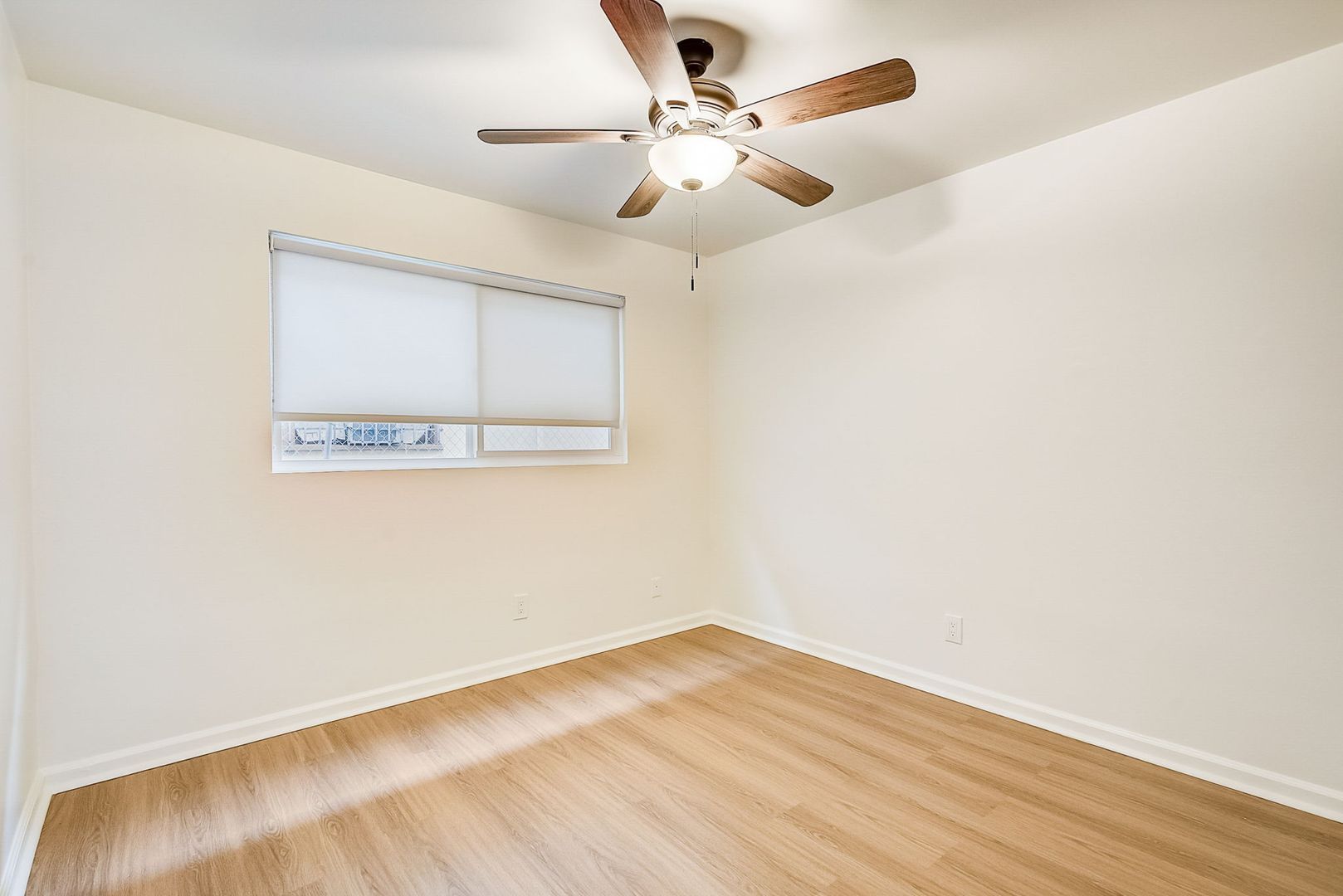 A vacant, neutral-walled bedroom with wood-look flooring, a window with closed blinds, and a central ceiling fan.