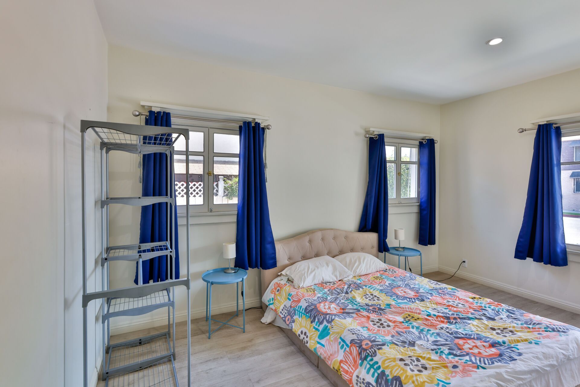 this image shows a bedroom with colorful bedsheet, three windows with blue curtains.