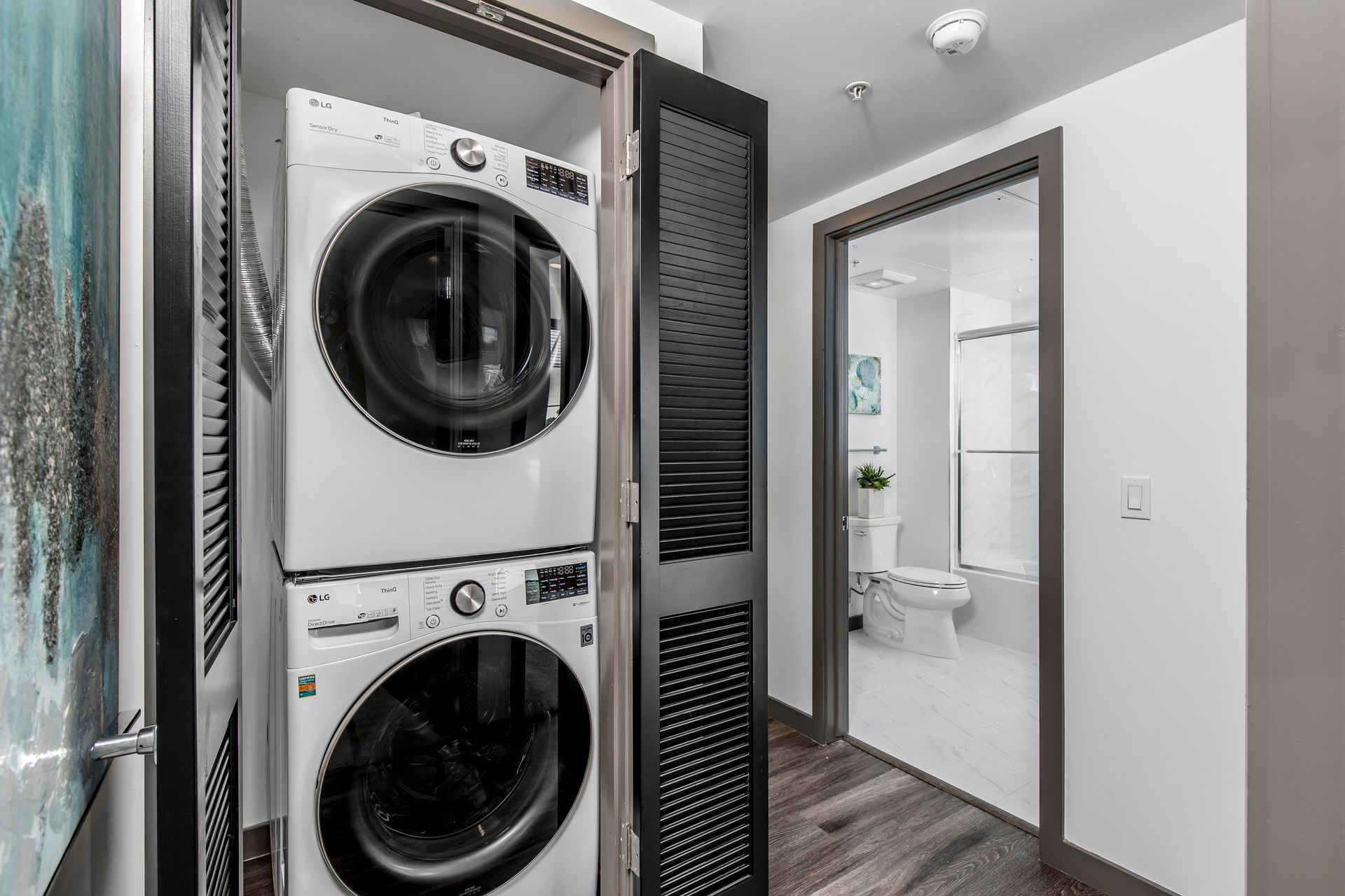 A white stacked washer and dryer in a closet with dark louvered doors, next to a doorway showing a bathroom.