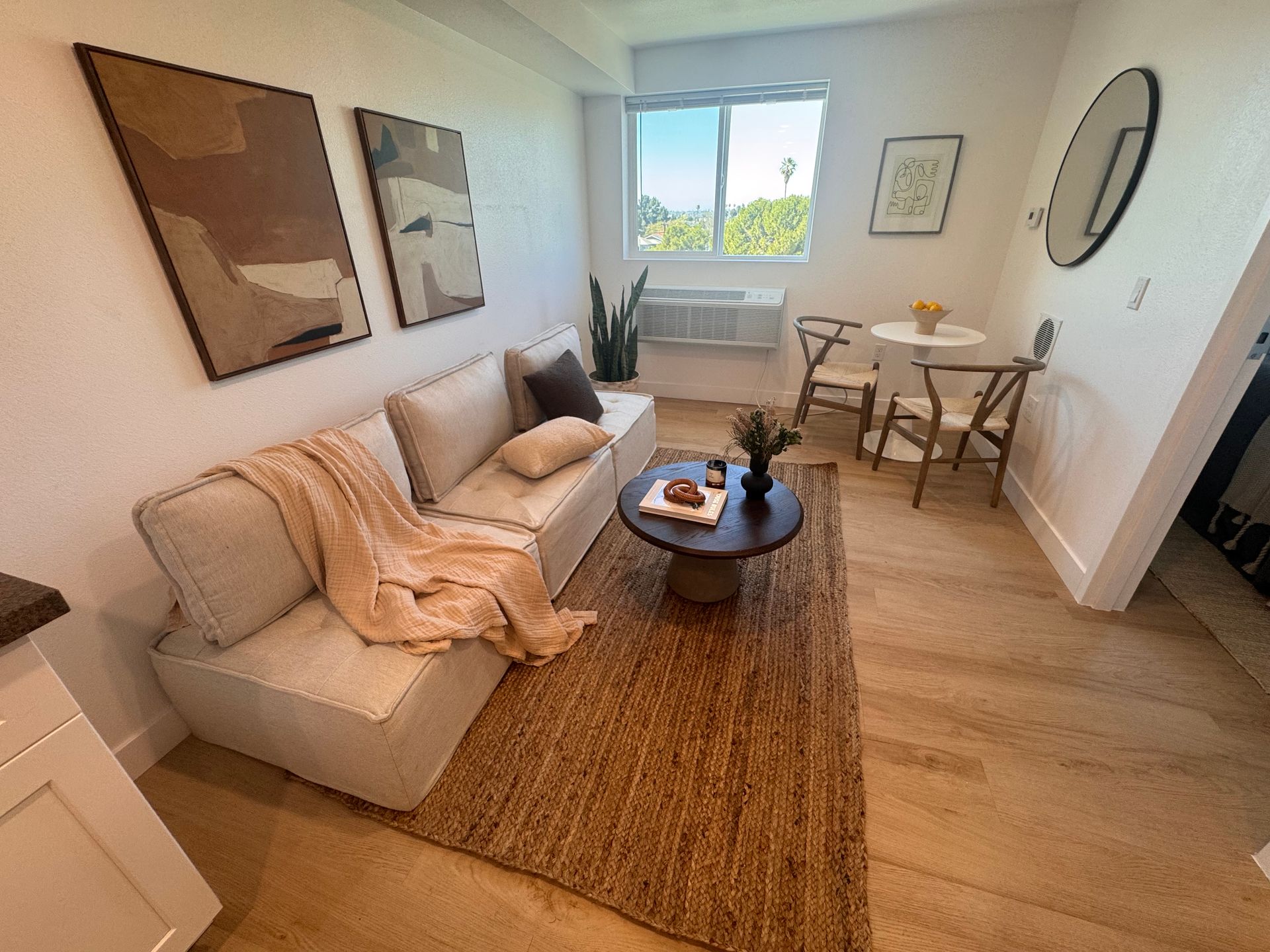 A modern living room with a beige sofa, jute rug, circular coffee table, and a small dining set near a bright window.