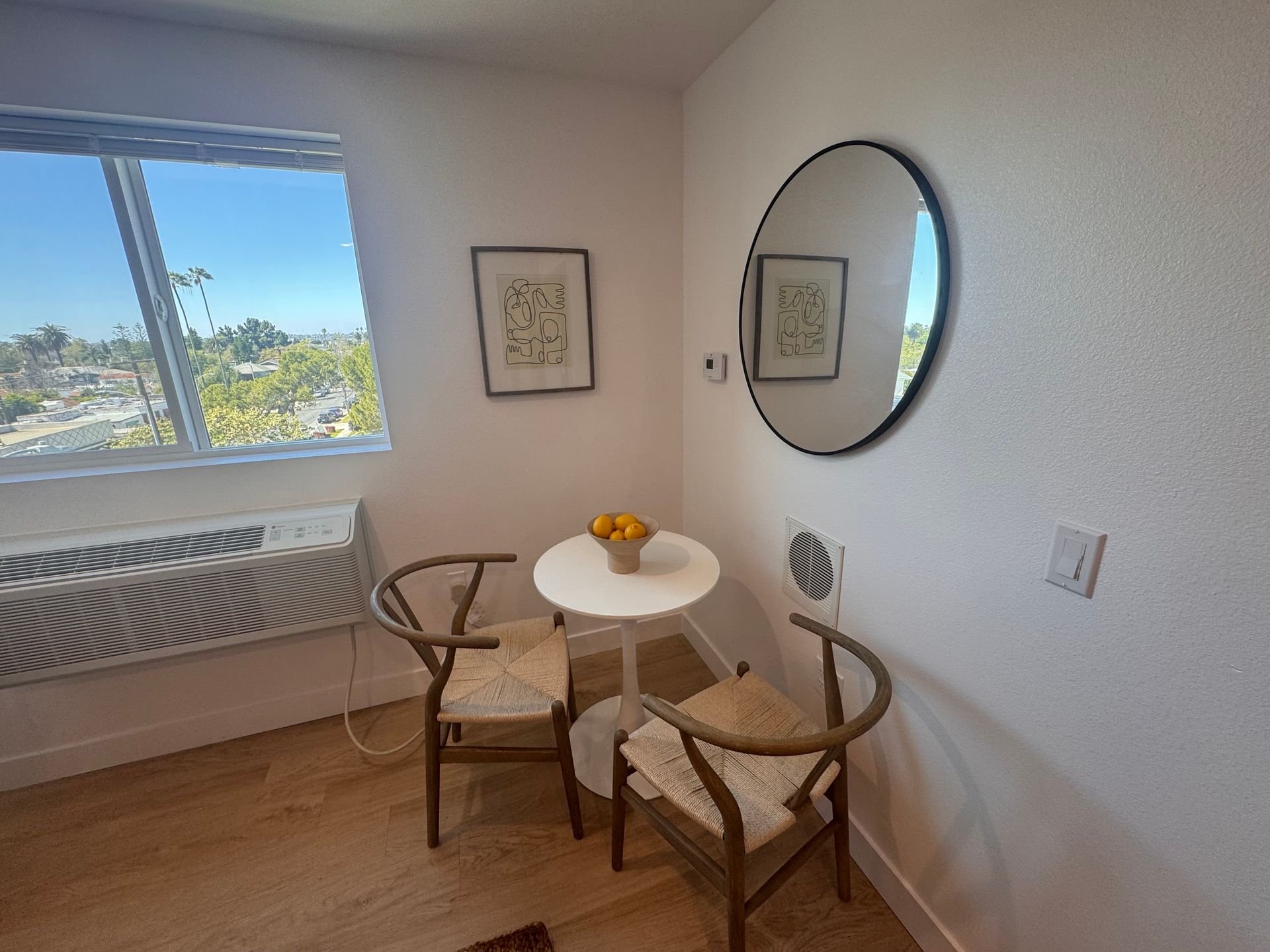 A small white bistro table with a bowl of lemons and two wooden chairs sits between a wall mirror and a room window.