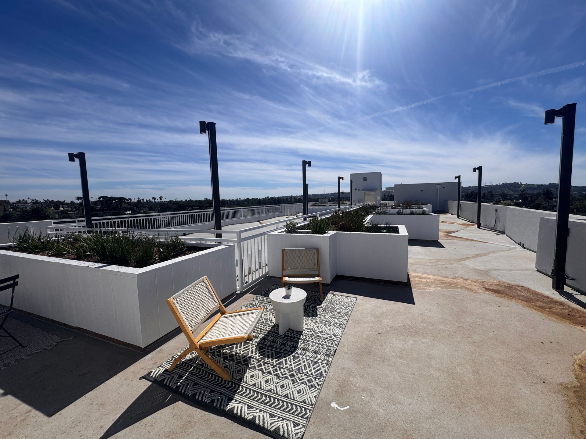 A rooftop deck with white planters, dark lampposts, and a small sitting area with two chairs on a patterned rug.