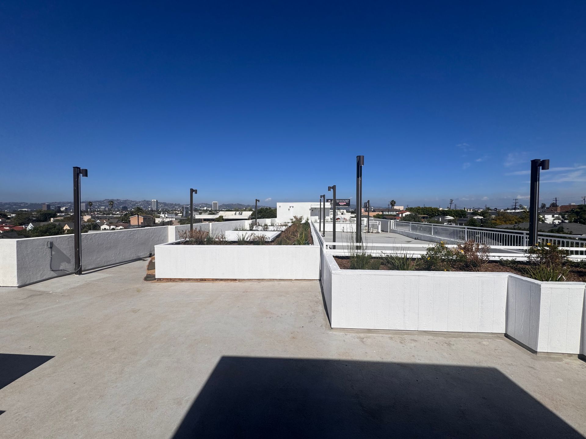 A rooftop patio with white concrete planter walls, outdoor lighting posts, and a city skyline view under a clear blue sky.