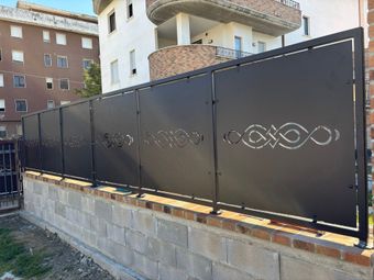 Recinzione in ferro nero con intagli decorativi lungo un muretto di pietra in una zona residenziale.