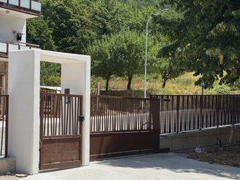 Cancello bianco e recinzione di legno marrone accanto a una casa, con alberi e una collina verde sullo sfondo.