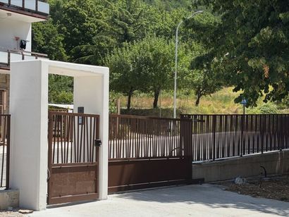 Cancello bianco e recinzione di legno marrone accanto a una casa, con alberi e una collina verde sullo sfondo.
