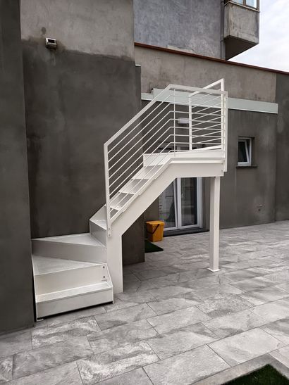 Scala esterna in metallo bianco addossata a un edificio grigio, in un cortile pavimentato.
