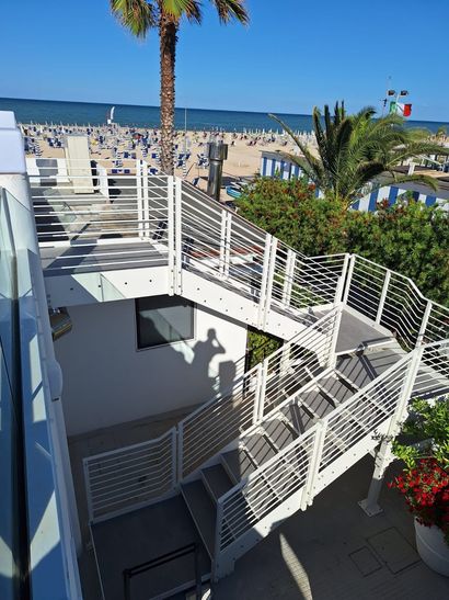 Scalinata e rampa bianche sul lungomare, con corrimano, accanto a una spiaggia sabbiosa e al mare, sotto un cielo azzurro e soleggiato