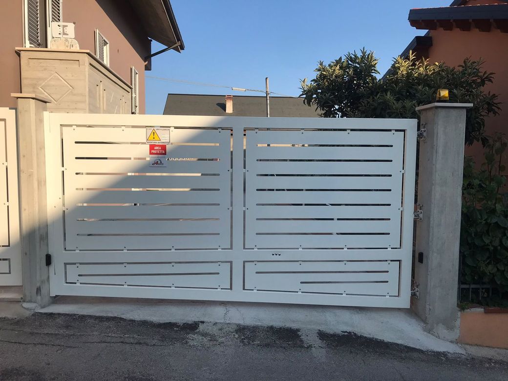 Cancello carrabile in metallo bianco davanti a una casa beige, con un piccolo cartello di avvertimento sul lato sinistro.