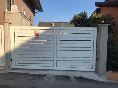 Cancello carrabile in metallo bianco davanti a una casa beige, con un piccolo cartello di avvertimento sul lato sinistro.