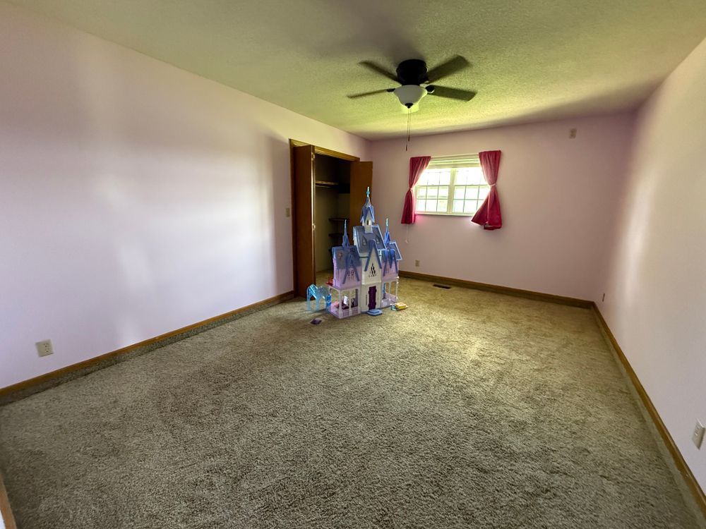 Empty carpeted room with ceiling fan, window with red curtains, and a small toy castle near the wall