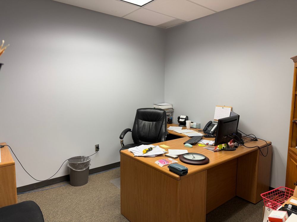 Empty office with desk, black chair, computer monitor, papers, and trash can against gray walls.