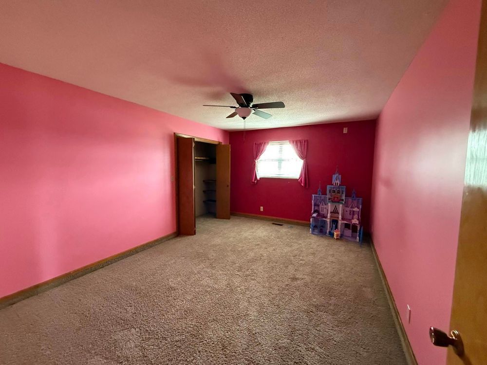 Empty pink bedroom with carpet, ceiling fan, window, and a small stack of toys in the corner