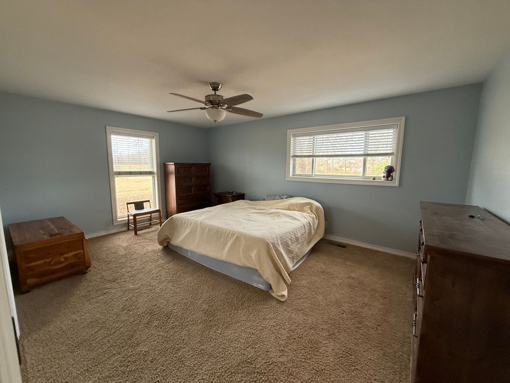 Spacious bedroom with a bed, dresser, chest, carpet, and two windows under a ceiling fan.