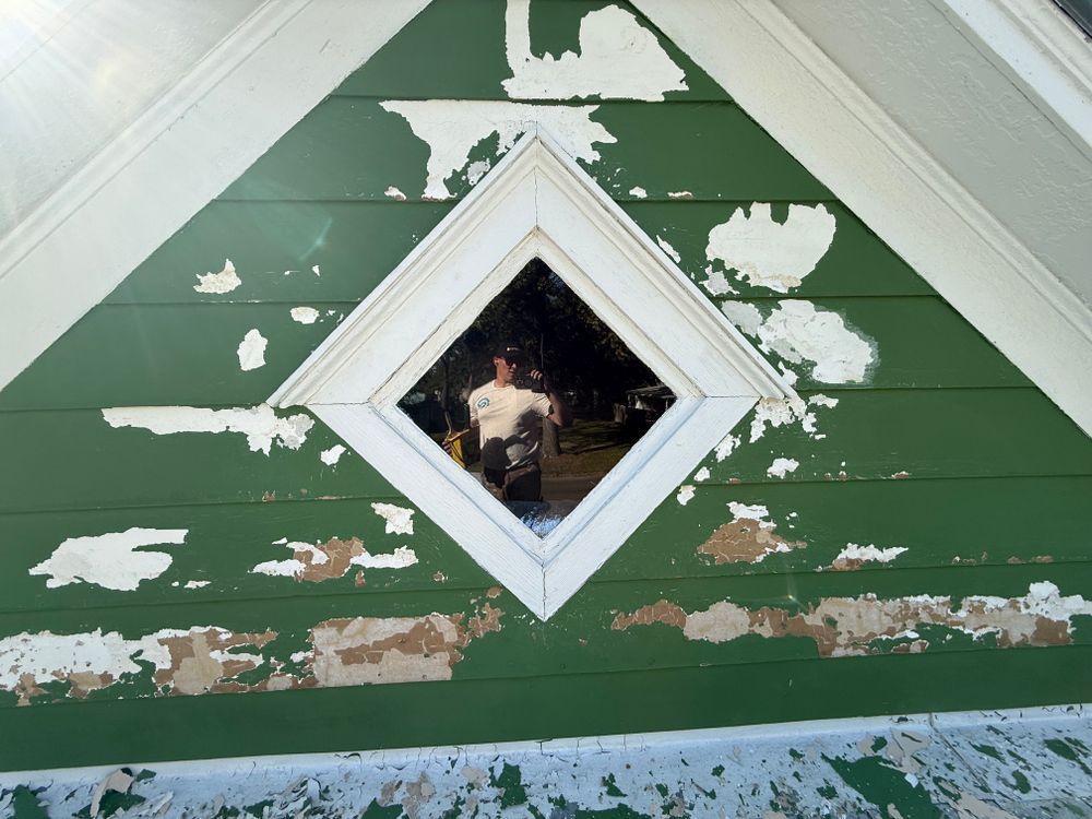 Peeling green siding with white trim and a diamond-shaped window on a house gable