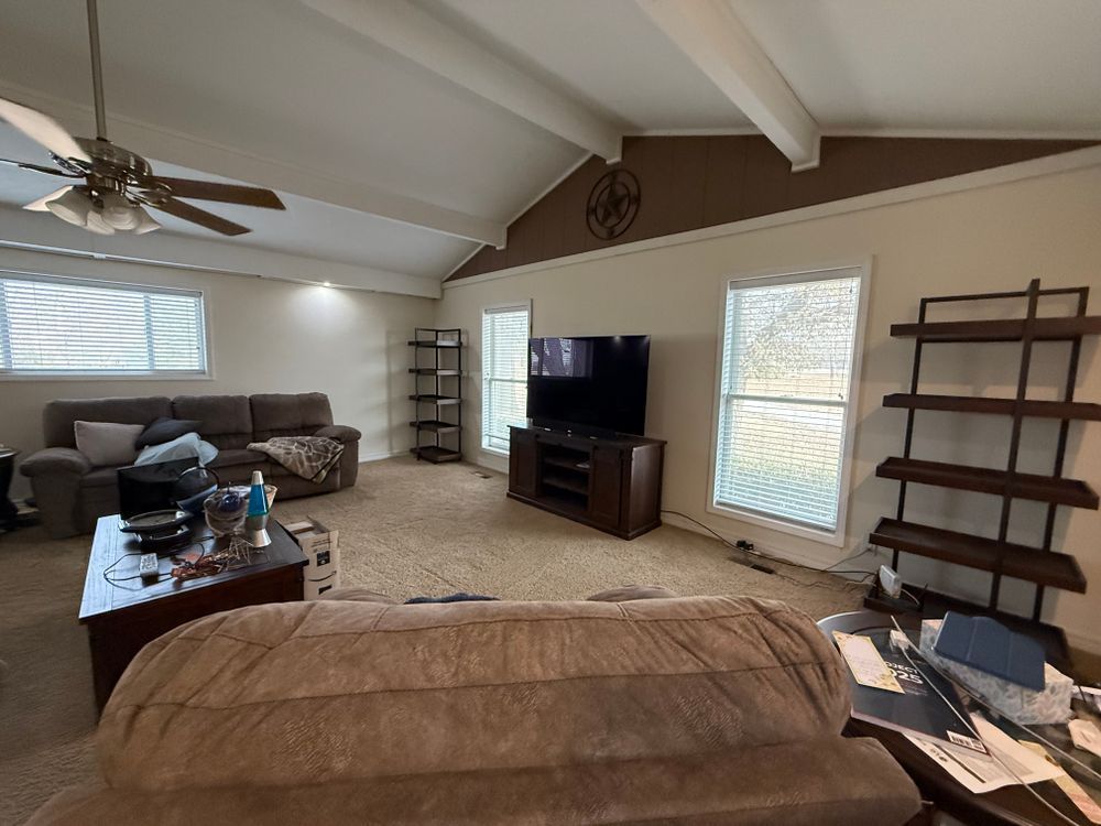 Spacious living room with beige carpet, ceiling fan, sectional sofa, TV, and empty wall shelves.