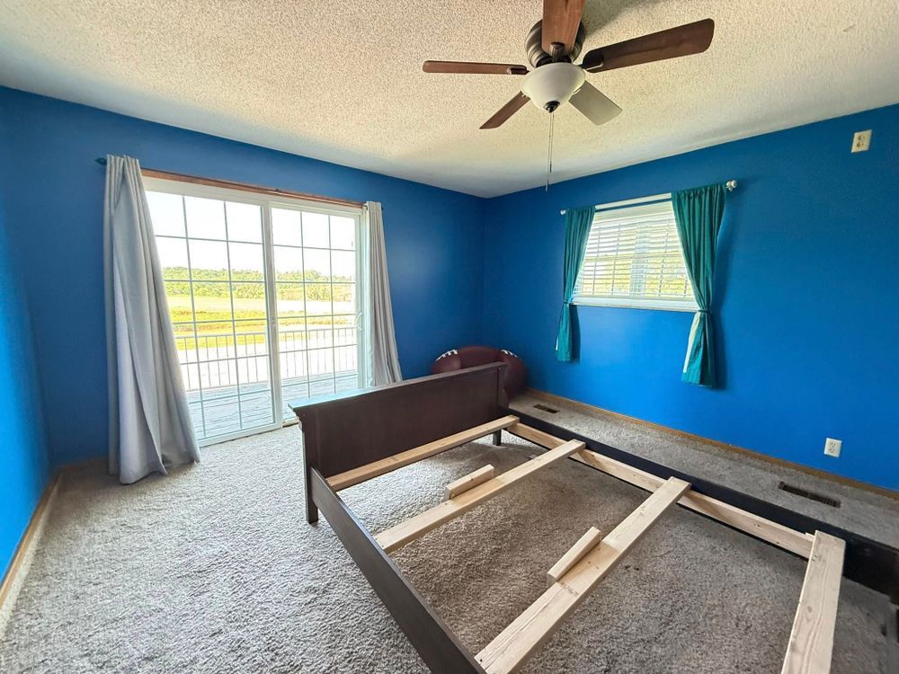 Blue bedroom with windows, ceiling fan, and a bed frame without a mattress