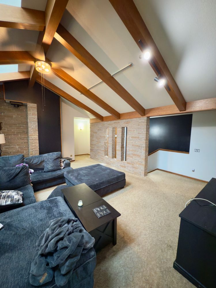 Modern living room with vaulted wood-beam ceiling, gray sectional, stone fireplace, and wall-mounted TV.