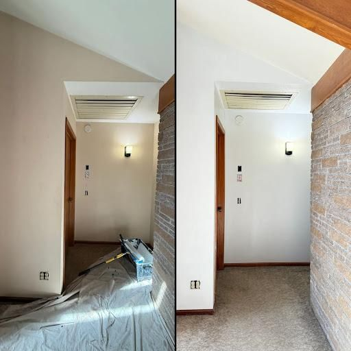 Empty hallway with beige walls, wall light, ceiling vent, and a brick accent wall; one side shows renovation debris.