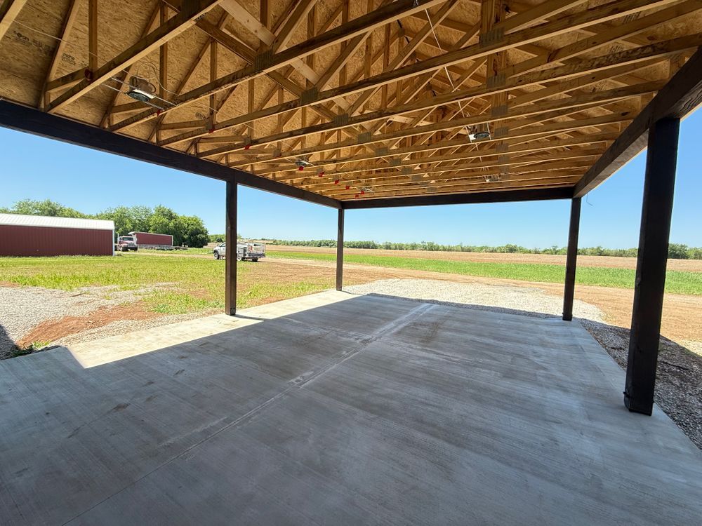 Open-sided pavilion with a concrete floor, wooden truss roof, and black support posts on a grassy rural site