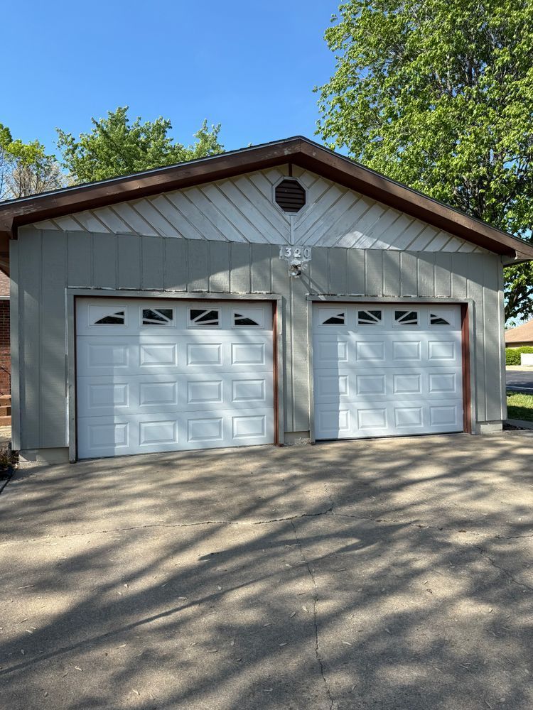 Two-car garage with white doors and gray siding, viewed from a driveway under trees.