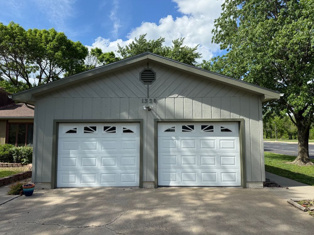 Two-car white garage with gabled roof and driveway, surrounded by trees and greenery