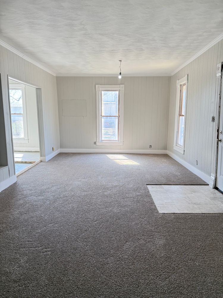 Empty carpeted room with white walls, tall windows, and a hanging light fixture.