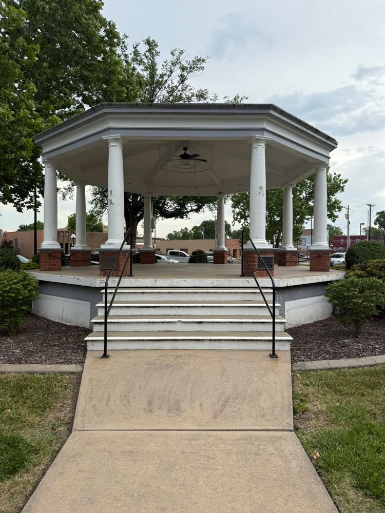 White gazebo with stairs and railings in a park, surrounded by trees and grass.