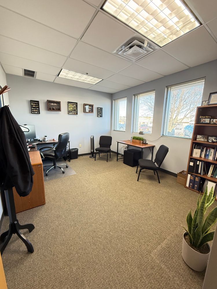 Small office with desks, chairs, framed wall art, bookshelves, and potted plants by bright windows.
