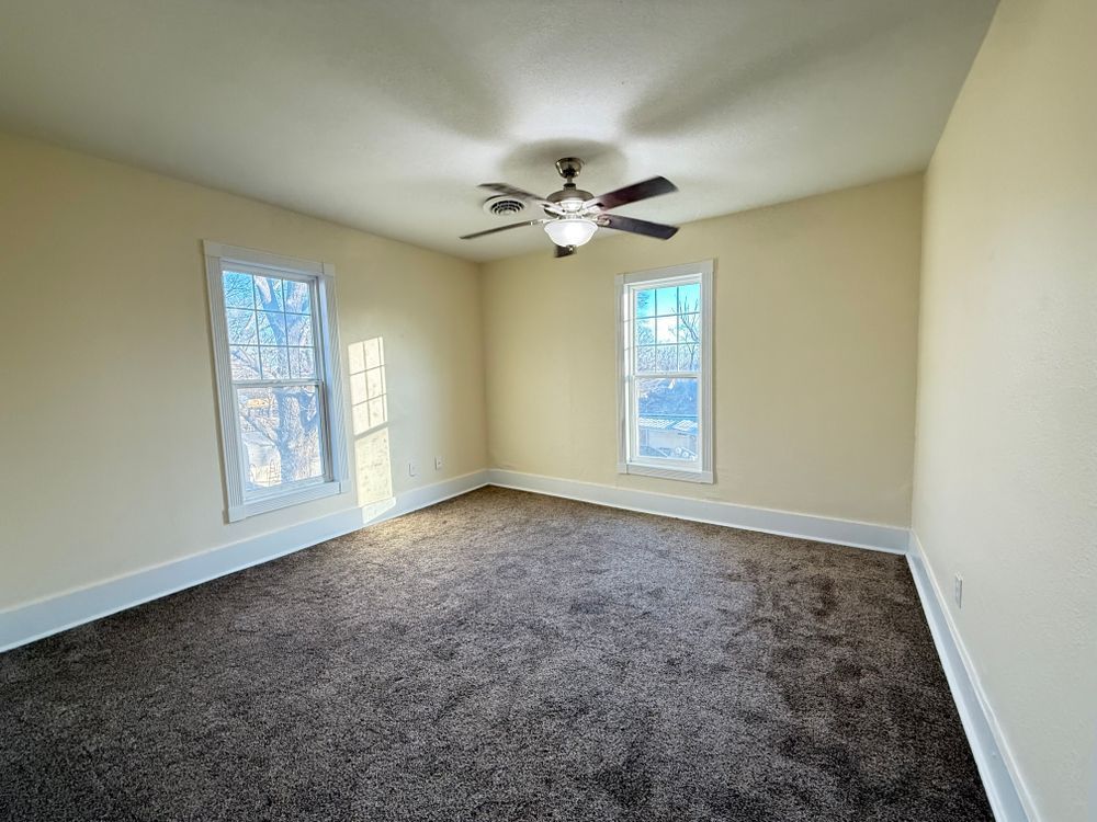 Empty beige bedroom with carpet, two windows, and a ceiling fan.