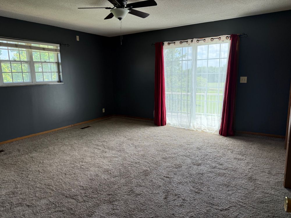 Empty living room with dark blue walls, beige carpet, ceiling fan, and sliding glass door with red curtains