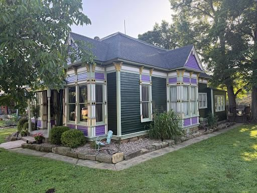 Small Victorian-style house with dark siding, purple trim, and a landscaped yard under trees