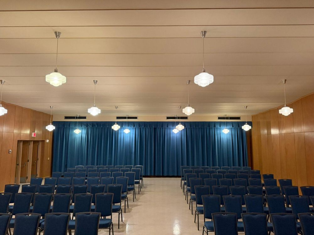 Empty auditorium with blue seats, wood-paneled walls, and blue stage curtains under ceiling lights