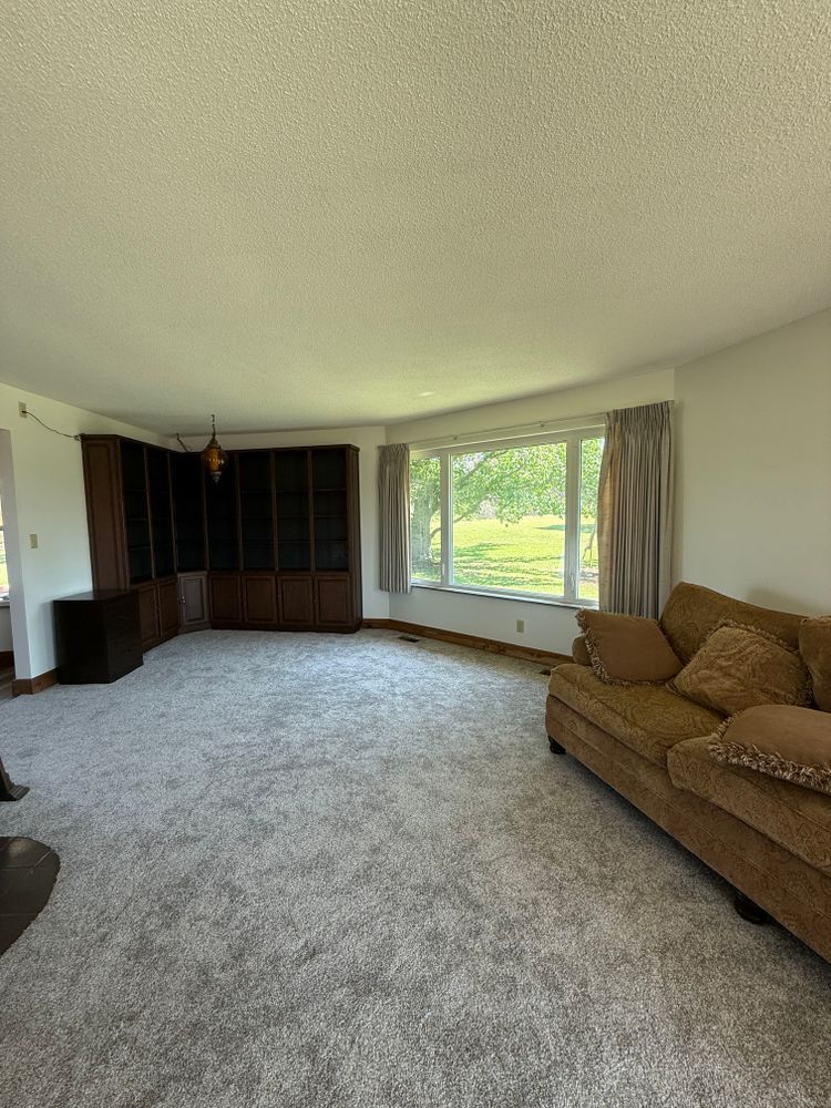 Empty living room with beige carpet, dark wood cabinets, and a large window with tan couch.