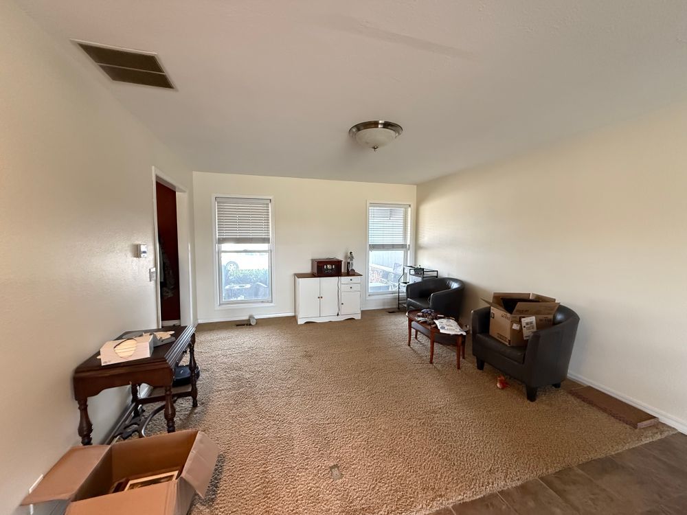 Empty living room with white walls, beige carpet, scattered furniture, and boxes near the window.