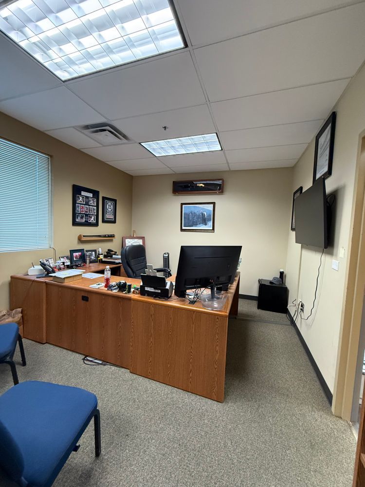 Small office with wooden desk, computer monitors, blue chair, and wall-mounted TV under fluorescent lights