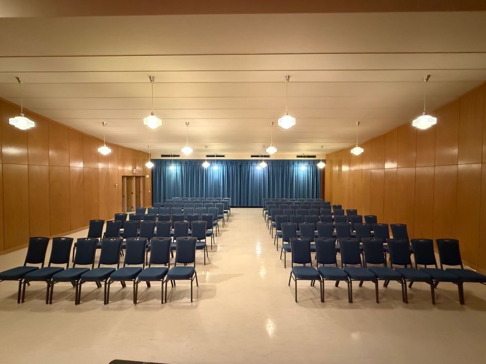 Empty conference room with rows of blue chairs facing a curtained stage and overhead lights